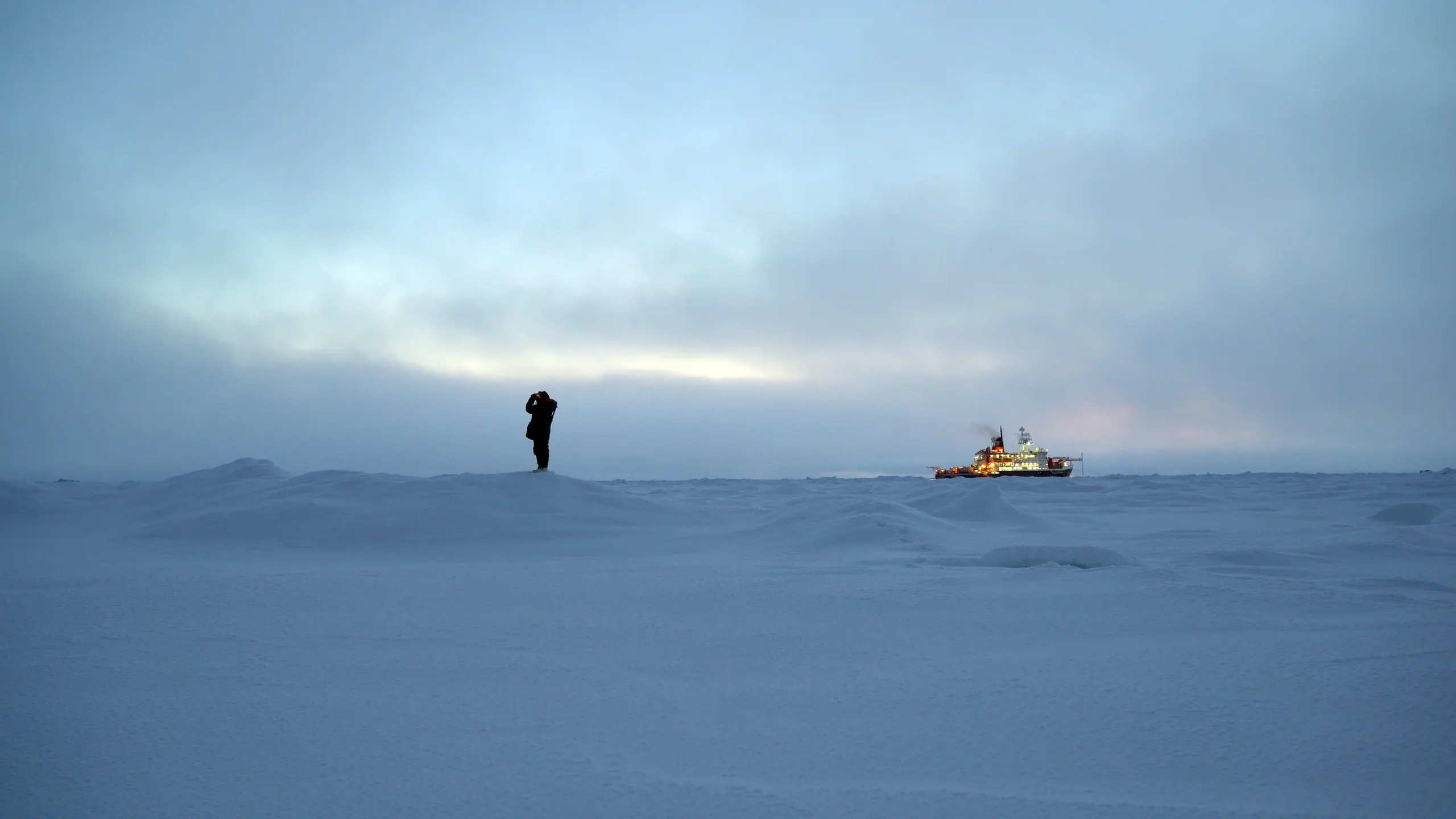 Photo of landscape with silhouette of figure holding binoculars and behind them in the distance a large well-lit work boat.