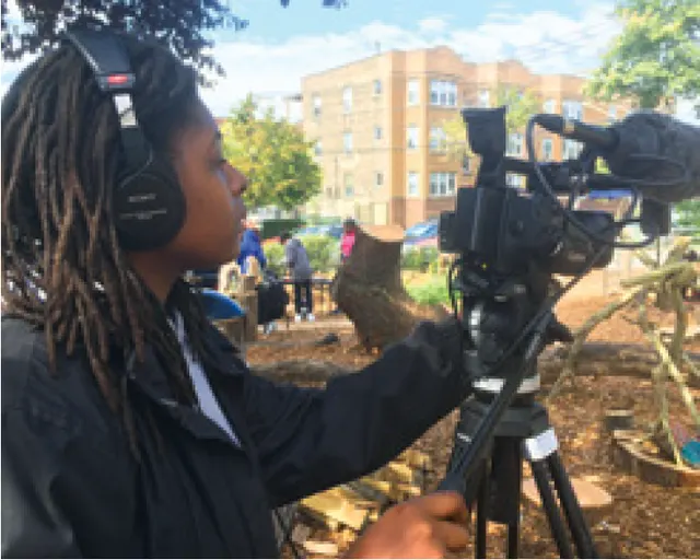 Black boy with dreads looks into a contemporary video camera with shotgun mic attached.