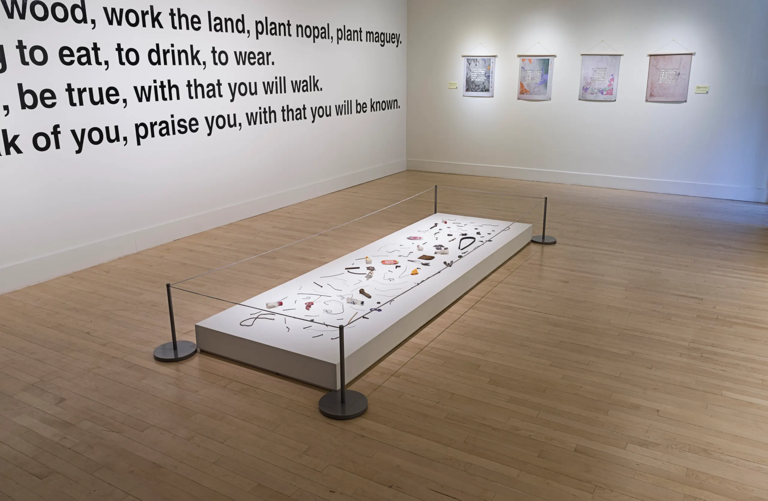 Photo of gallery with large black writing on white wall and a short table like pedestal on the floor exhibiting many small artifacts. Behind the floor pedestal on the wall hang four printed works.