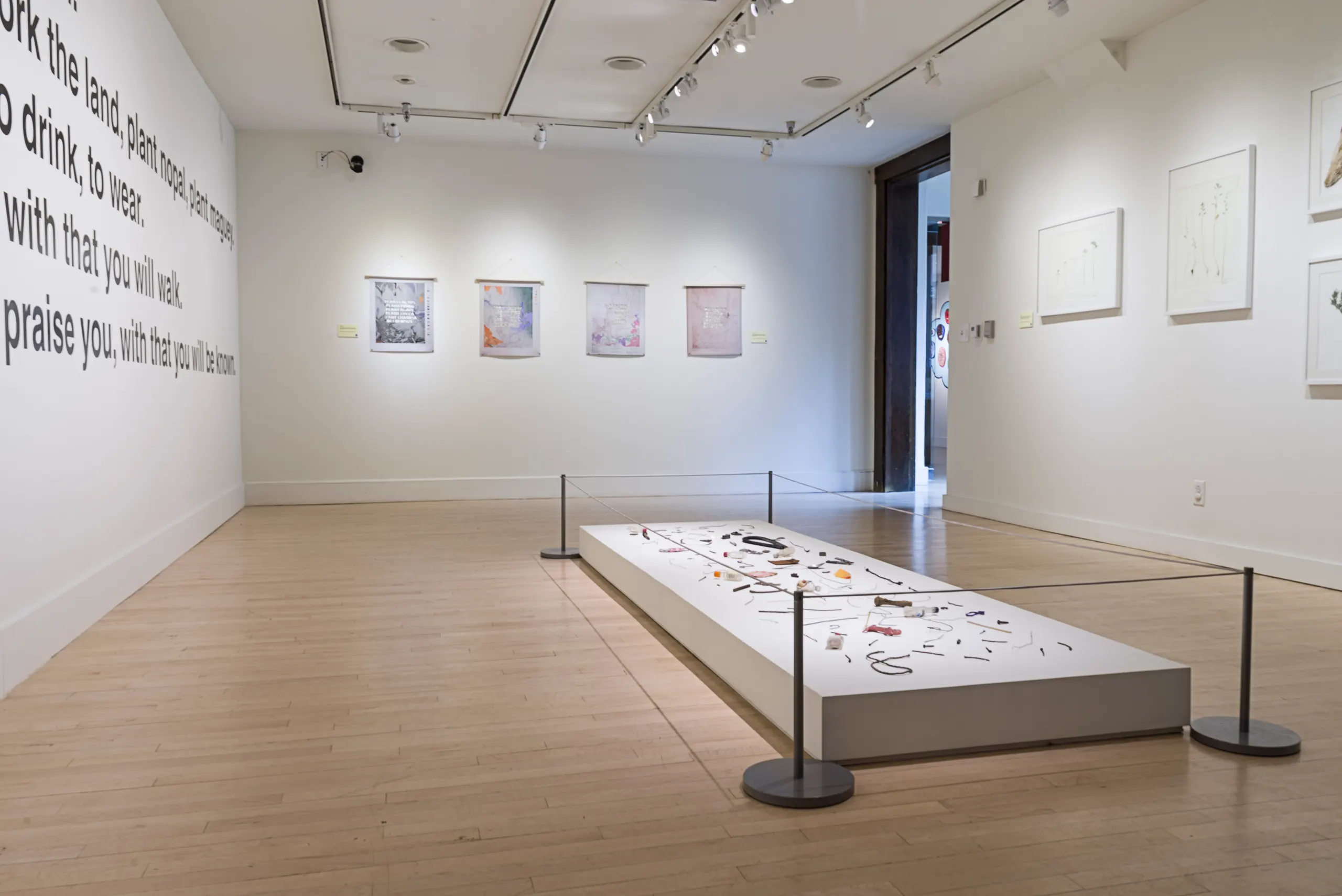 Photo of gallery with large black writing on white wall and a short table like pedestal on the floor exhibiting many small artifacts. Behind the floor pedestal on the wall hang four printed works.