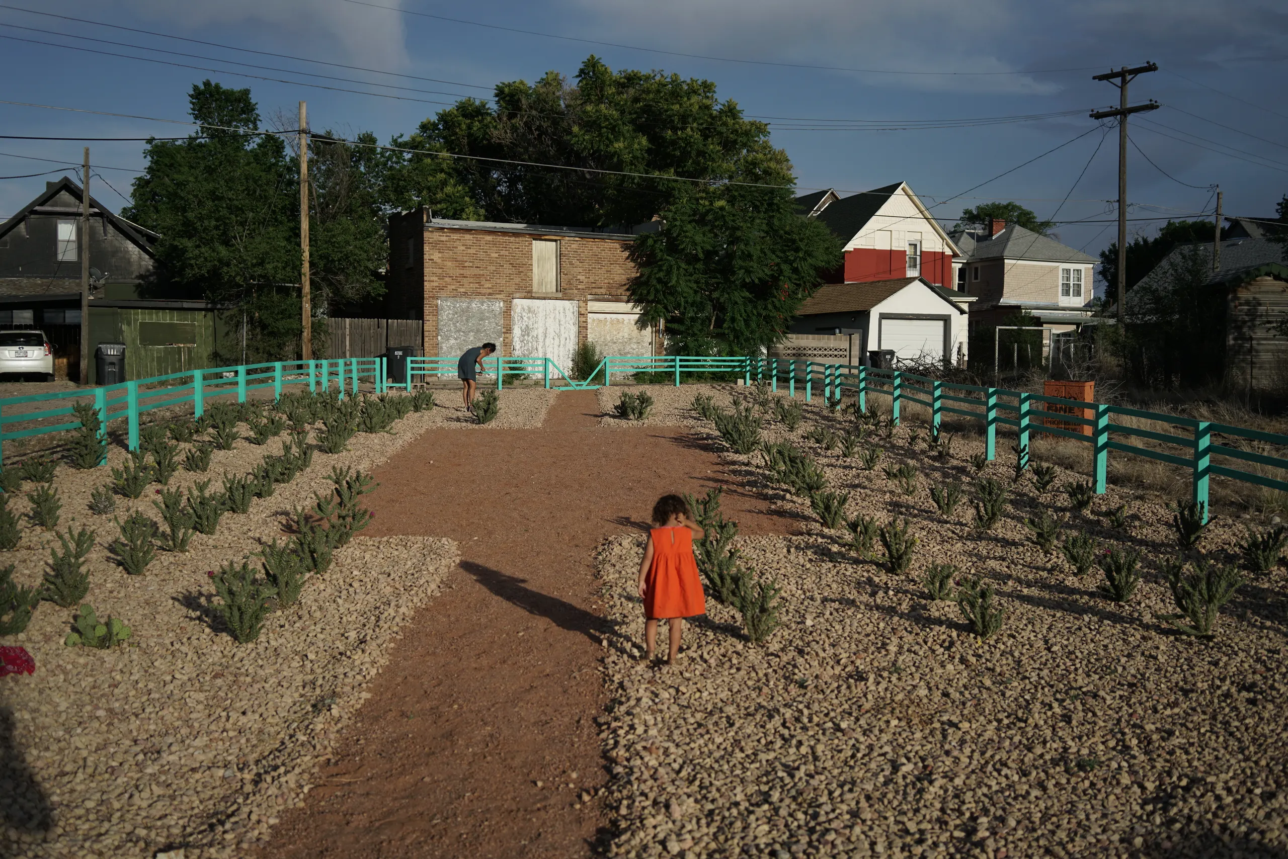 Photo of cacti garden with stone covering and blue fence. In the garden are a girl in a red dress in the foreground and woman in a blue jumper in the background.