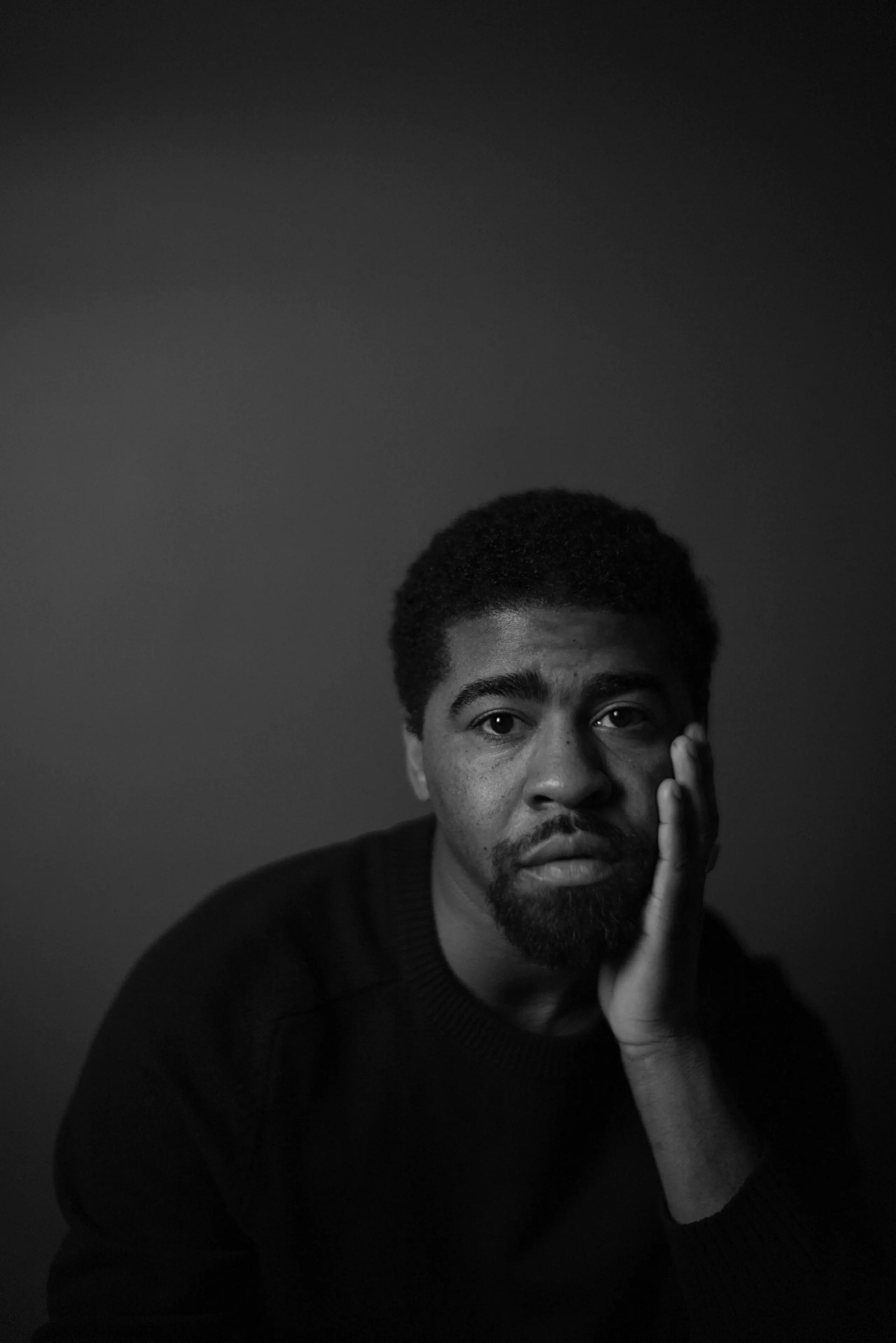 dark photo portrait of Devin, a black man with with a short dark hair and a gotee, looking at the camera with the left hand on his face.