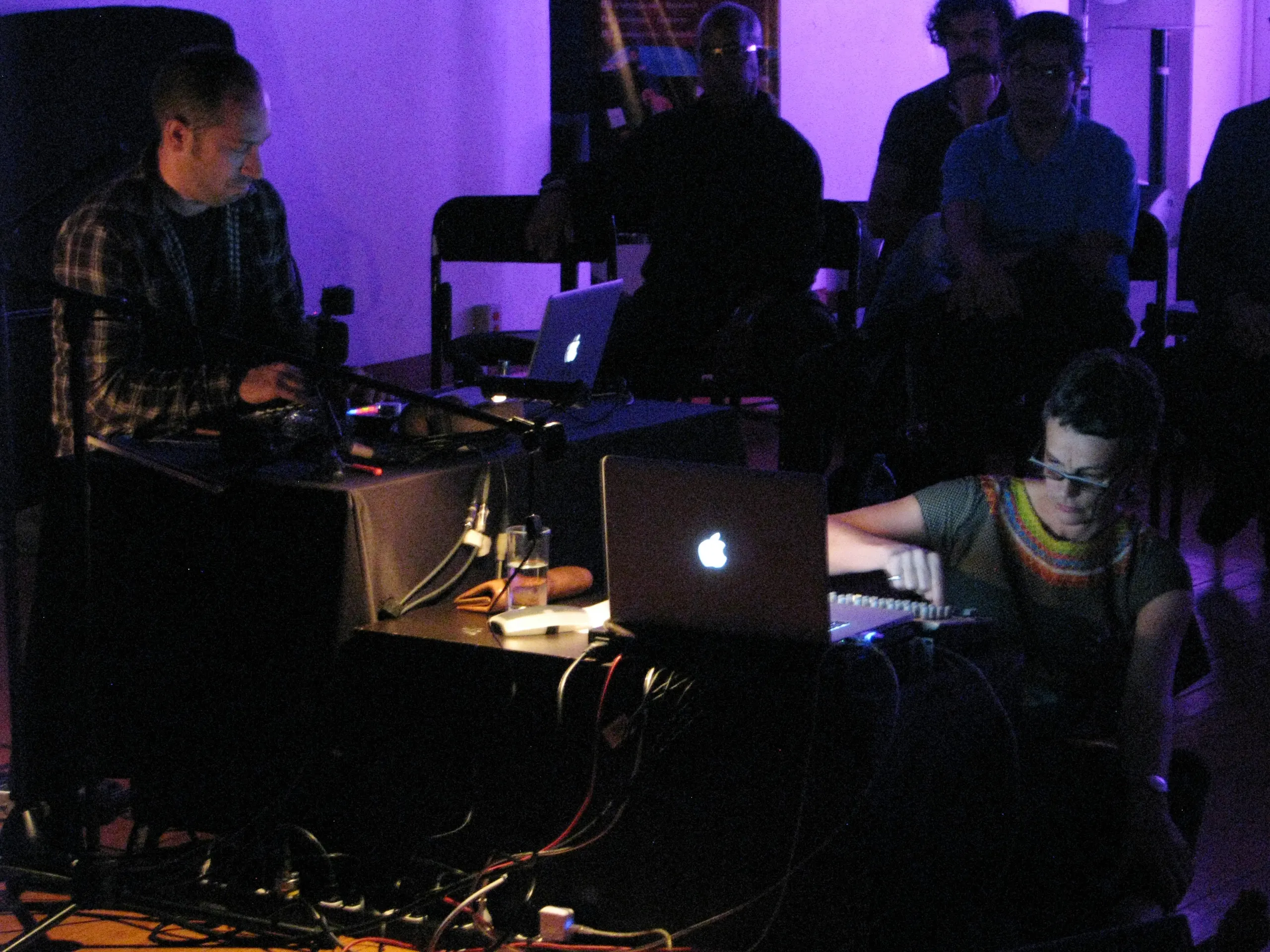 Photo of Schimana looking towards her laptop and turning knobs on mixing desk with another laptop player to the left and the audience behind them both.
