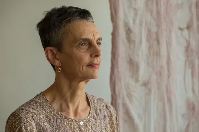 Photo portrait of Elisabeth Schimana with short black hair and an earth-toned sweater and ear rings and looking to the right.