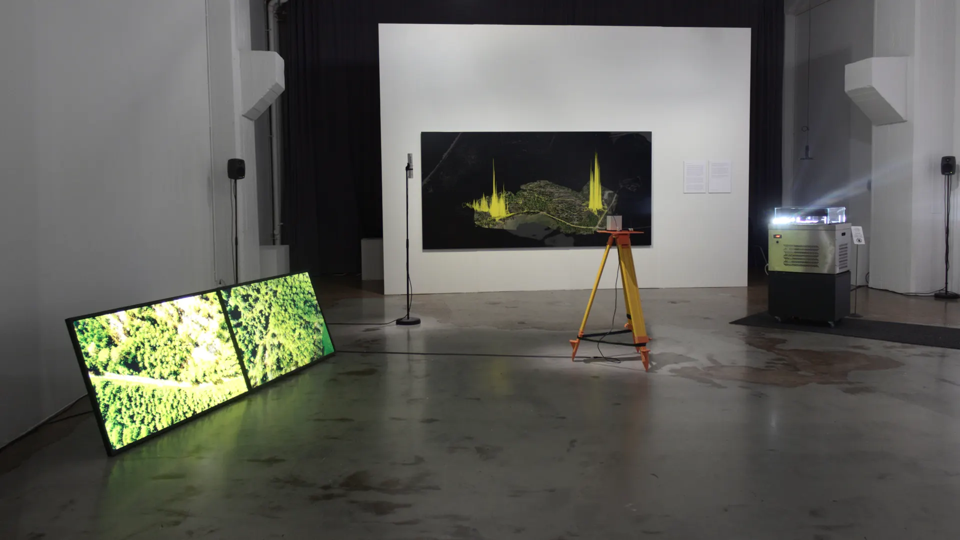 Photo of a the installation "Landscape Machines" by Erich Berger, including multiple video screens on the floor in a large open space with a hanging picture of a landscape map with spectral graphics overlaid.