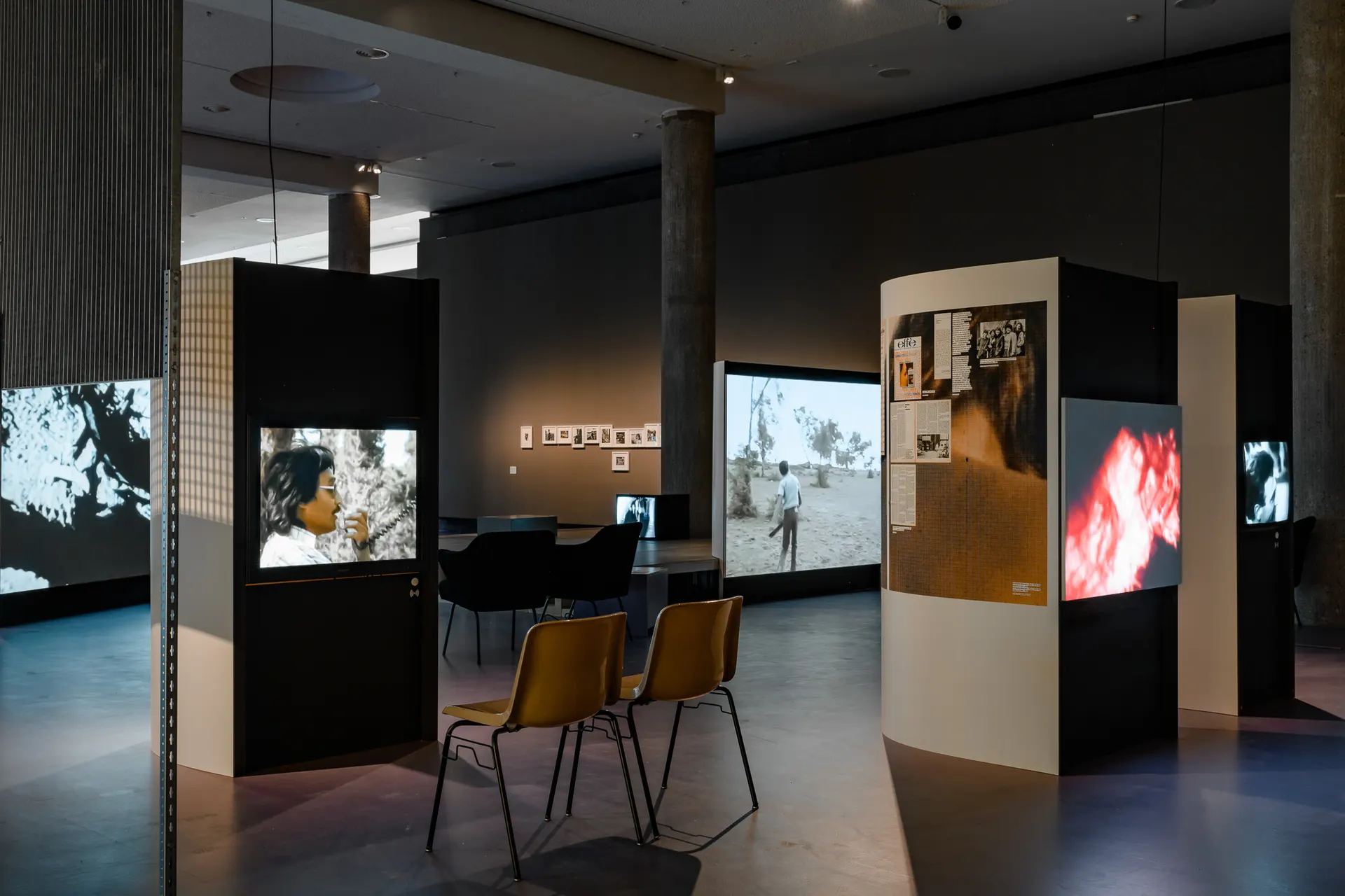 Photo of dark exhibition room with tall ceilings and many screen based works on display. In the foreground are two chairs facing a screen showing a man with dark hair speaking into a walkie-talkie like device.