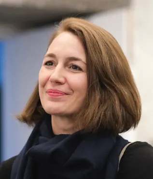 Photo portrait of Erika Balsom in black coat with shoulder-length brown hair, smiling and looking to the left of view.