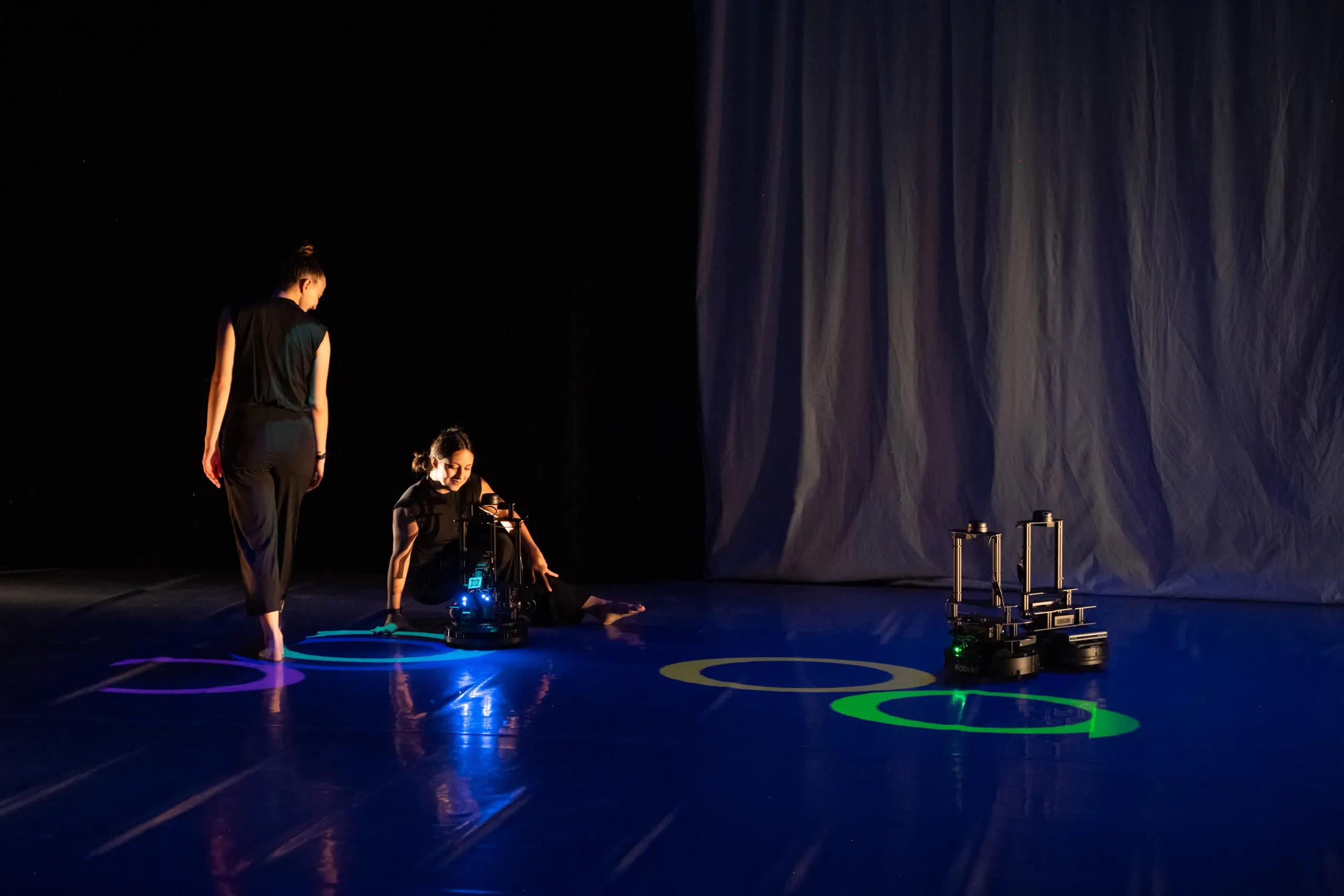 Color photo of three robots and two humans in a theater with a blueish floor