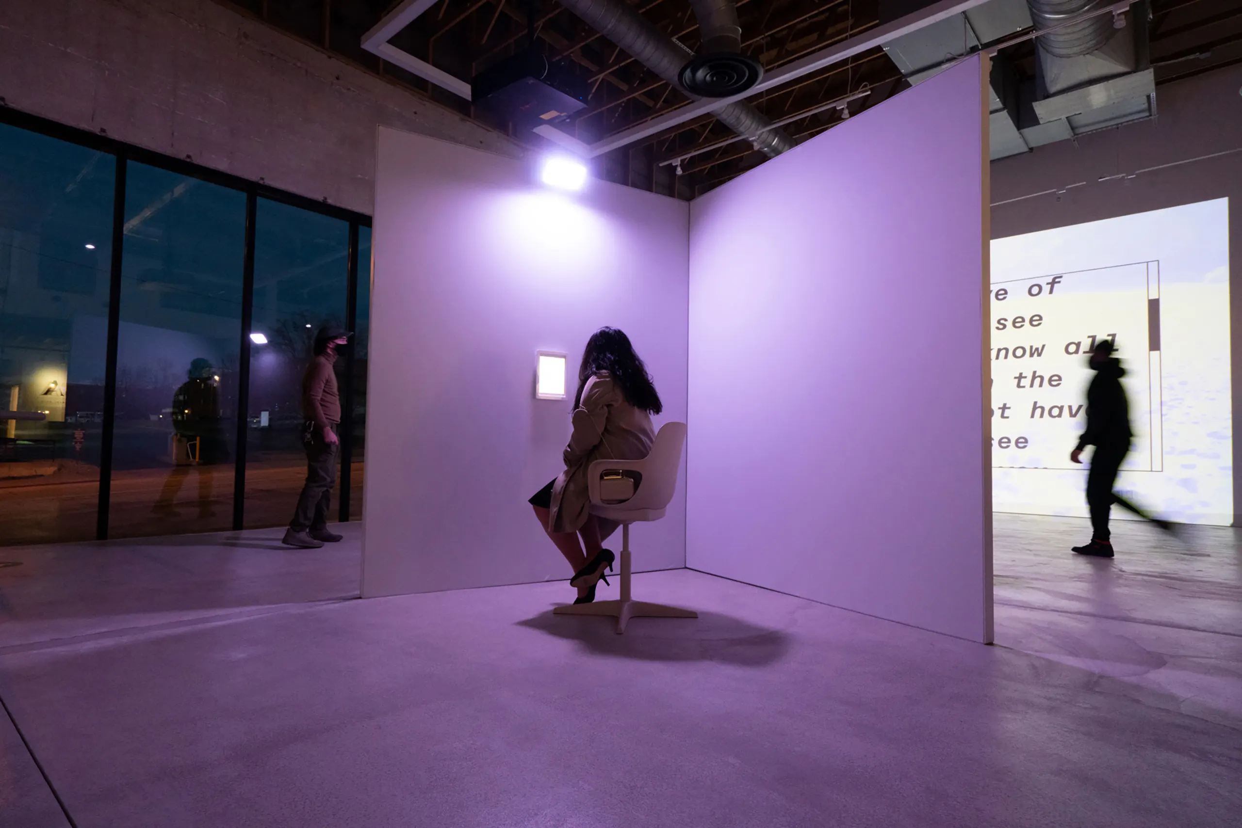 View of woman from behind with dark hair, sitting in a chair in a purpleish room looking at a glowing tablet on the wall