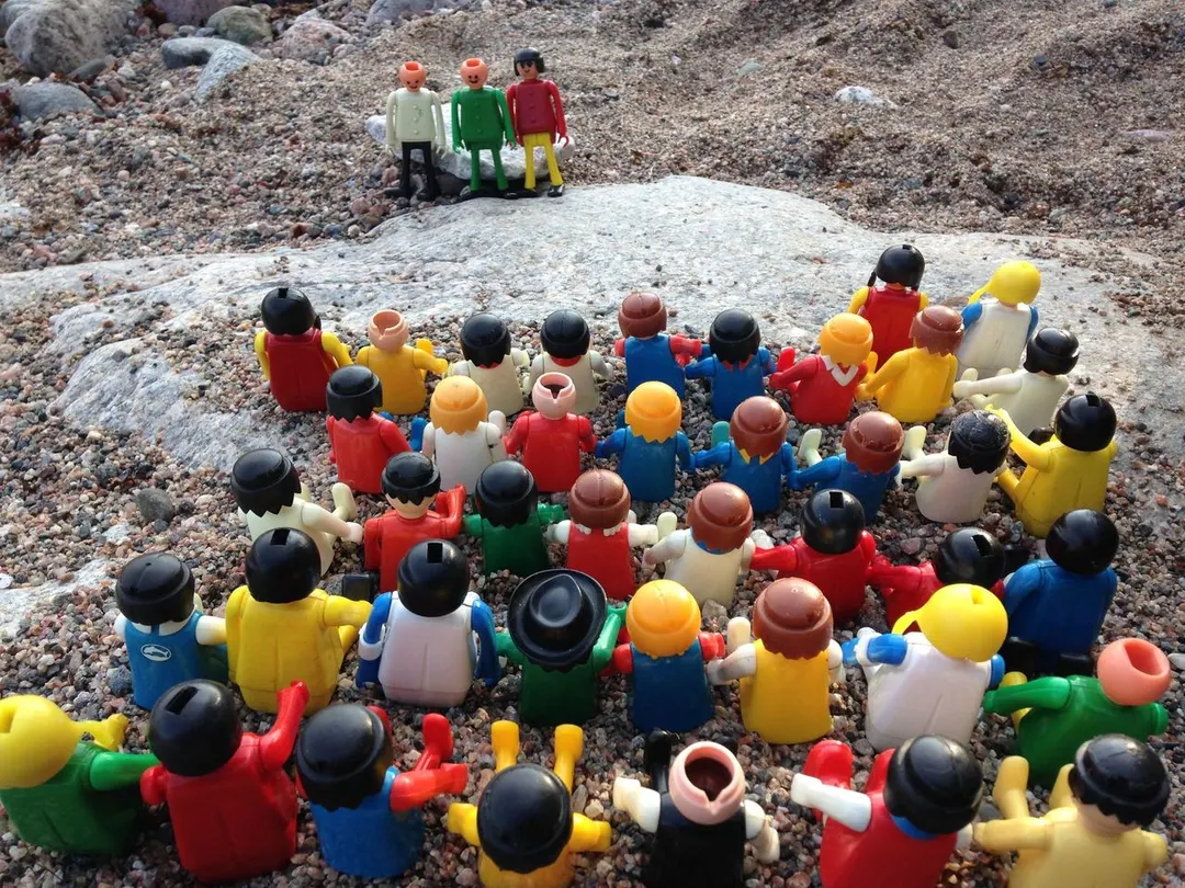 A group of 50 small colorful plastic toy figures take part in a ceremony outside in a Finnish archipelago landscape by a rocky shore.