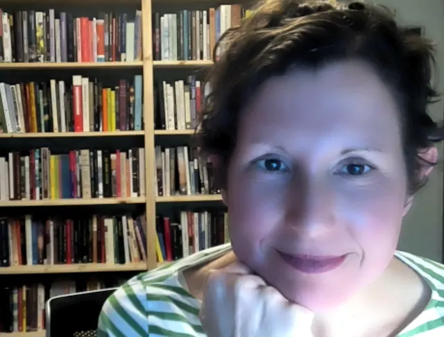 Photo portrait of Shannon, a white woman with dark hair and eyes, framed almost entirely on the face. She is looking straight at the camera with a soft smile, resting her face on her right hand under her chin. She is wearing a green and white striped shirt. In the background, we see a full bookshelf.