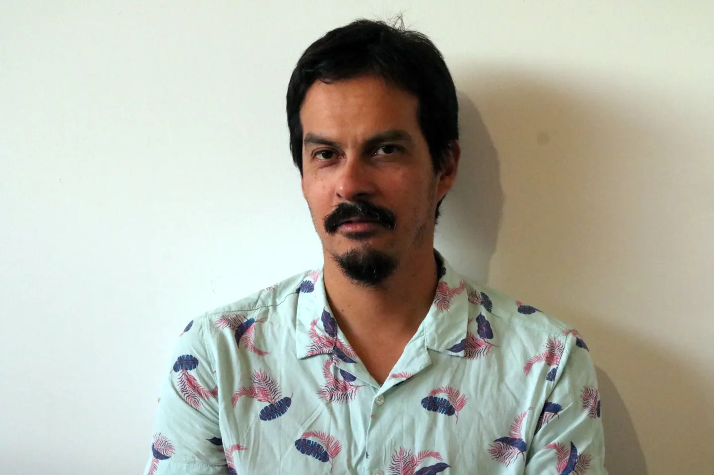 photo portrait of Fernando, dark skinned and dark haired man with mustache and goatee looking directly at camera with blue-green shirt covered in spaced patterns of blue and red overlapping feathers.